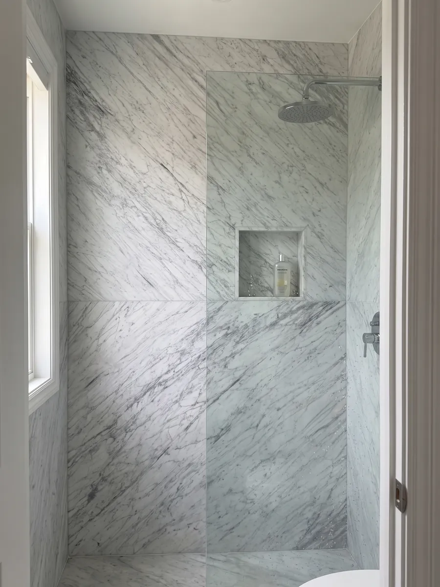 Shower clad in full slabs of white Carrara marble: grayish white with a fine, diffuse gray pattern spread across the whole surface