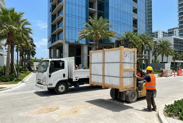A forklift unloads a crate of marble slabs from a truck in front of a high-rise under construction