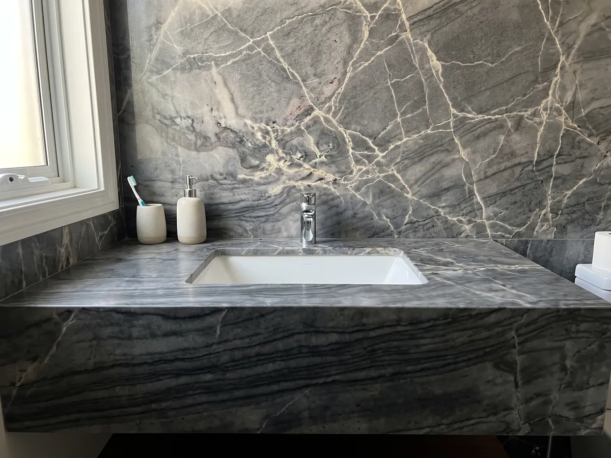 Vanity top and bathroom wall in London Grey quartzite with a white sink and chrome faucets: charcoal grey with a network of cream veins and dark horizontal waves