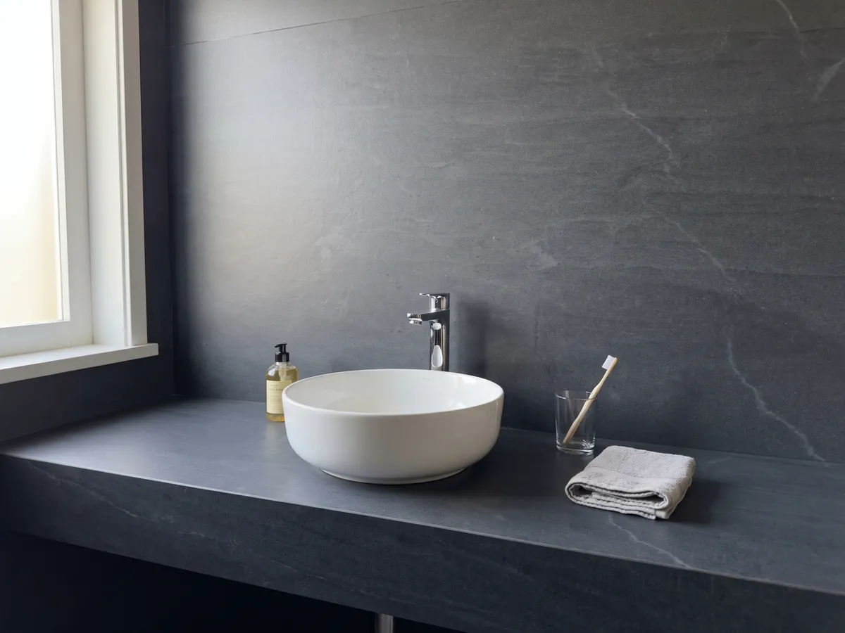 Vanity top and bathroom wall in gray Pietra del Cardoso quartzite with a white sink and chrome faucets: uniform dark slate gray, no pattern