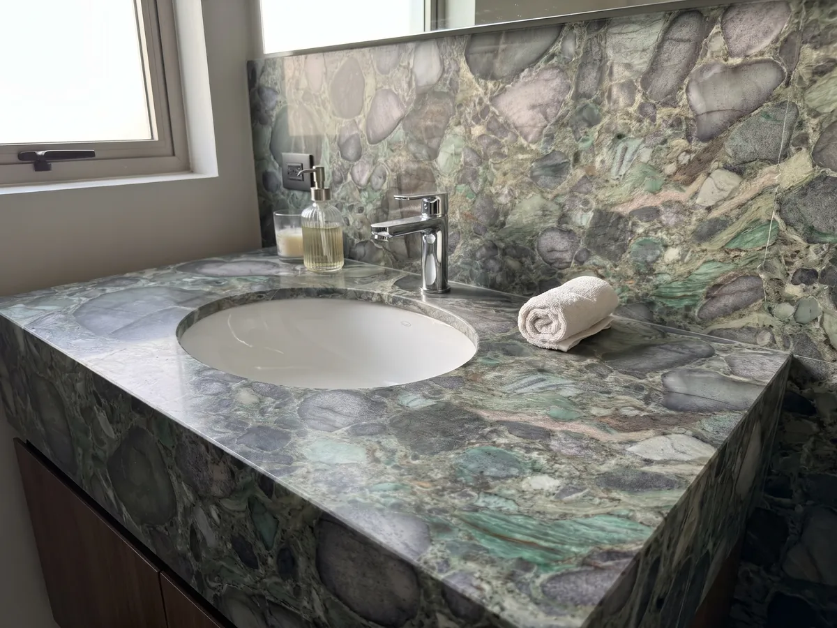 Bathroom vanity top and wall in Palladium Green quartzite with a white sink and chrome faucet: rounded violet-gray clasts with a green matrix and emerald glints