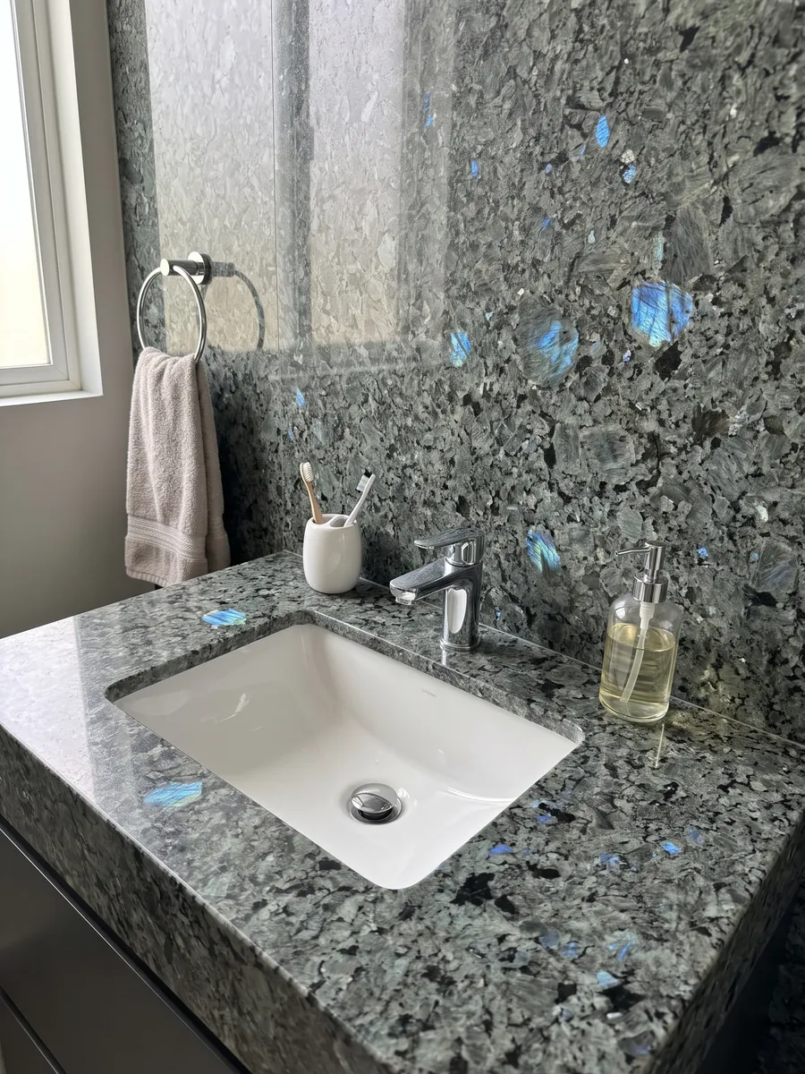 Vanity top and bathroom wall in Labradorite granite with a white sink and chrome fixtures: large greenish-gray crystals with iridescent blue flashes