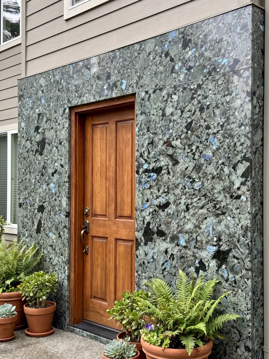 House facade clad in Labradorite granite next to a wood door: large greenish-gray crystals with iridescent blue flashes
