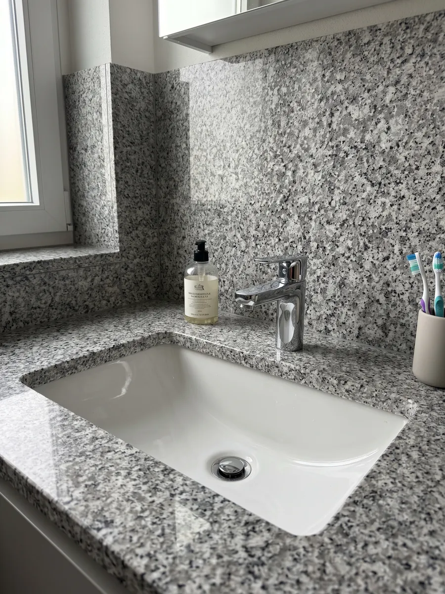 Vanity top and bathroom wall in Sardo Grigio granite with a white sink and chrome faucets: light gray salt and pepper with a medium grain
