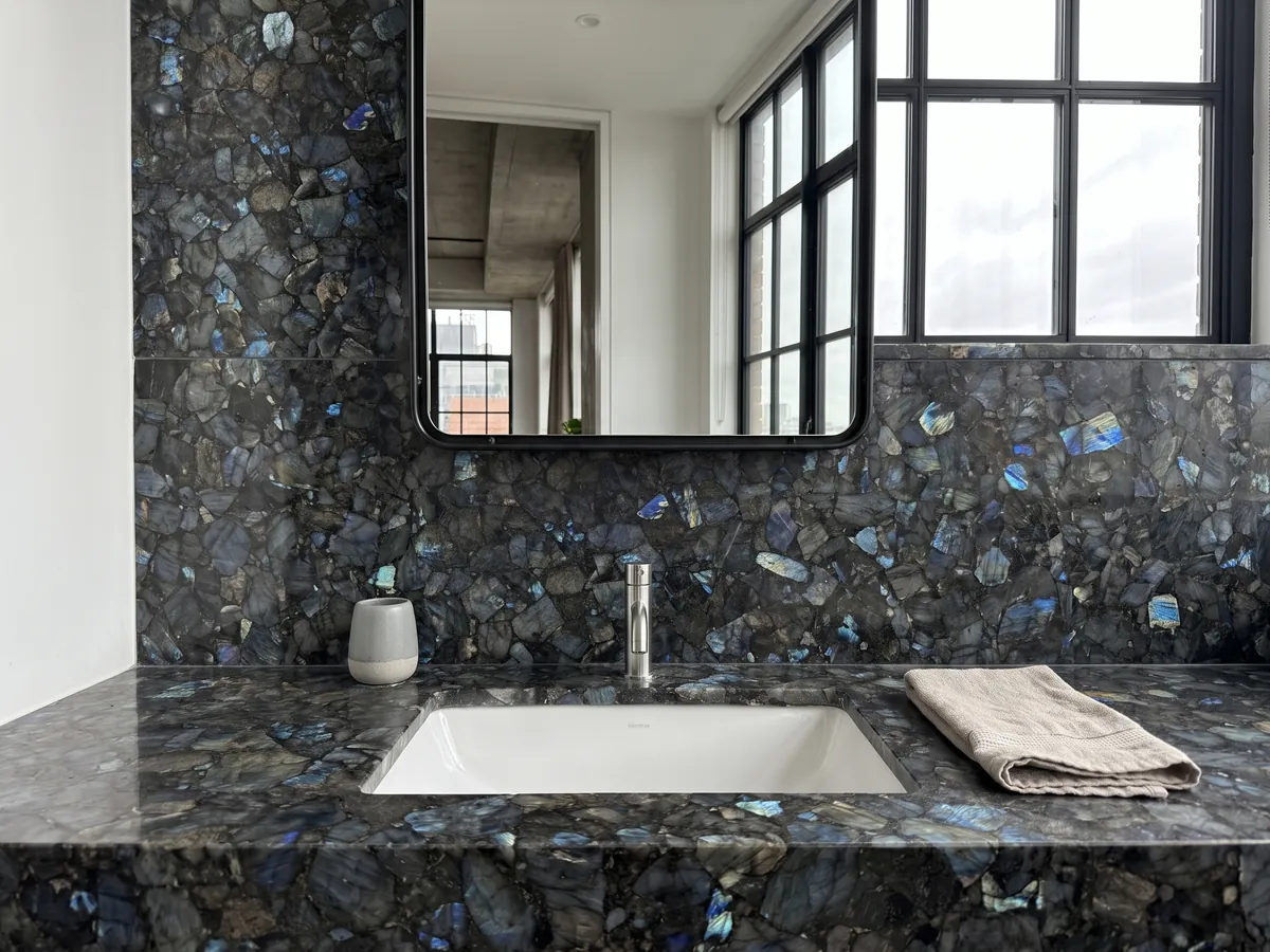 Vanity top and bathroom wall in labradorite with a white sink and chrome fixtures: dark gray pieces with iridescent blue flashes spread across