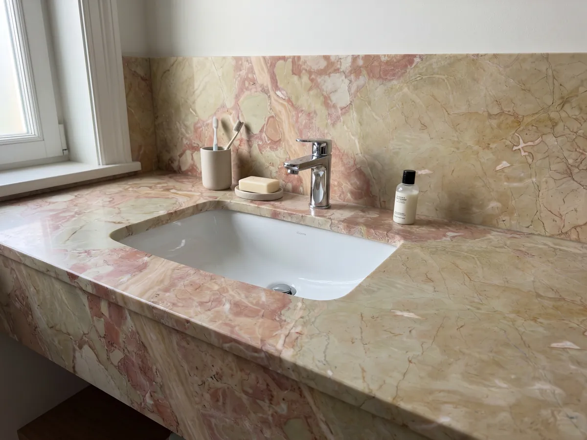 Bathroom vanity top and wall in beige Breccia D'Aragona marble with a white sink and chrome fixtures: large greenish beige fragments with coral pink patches along the left edge