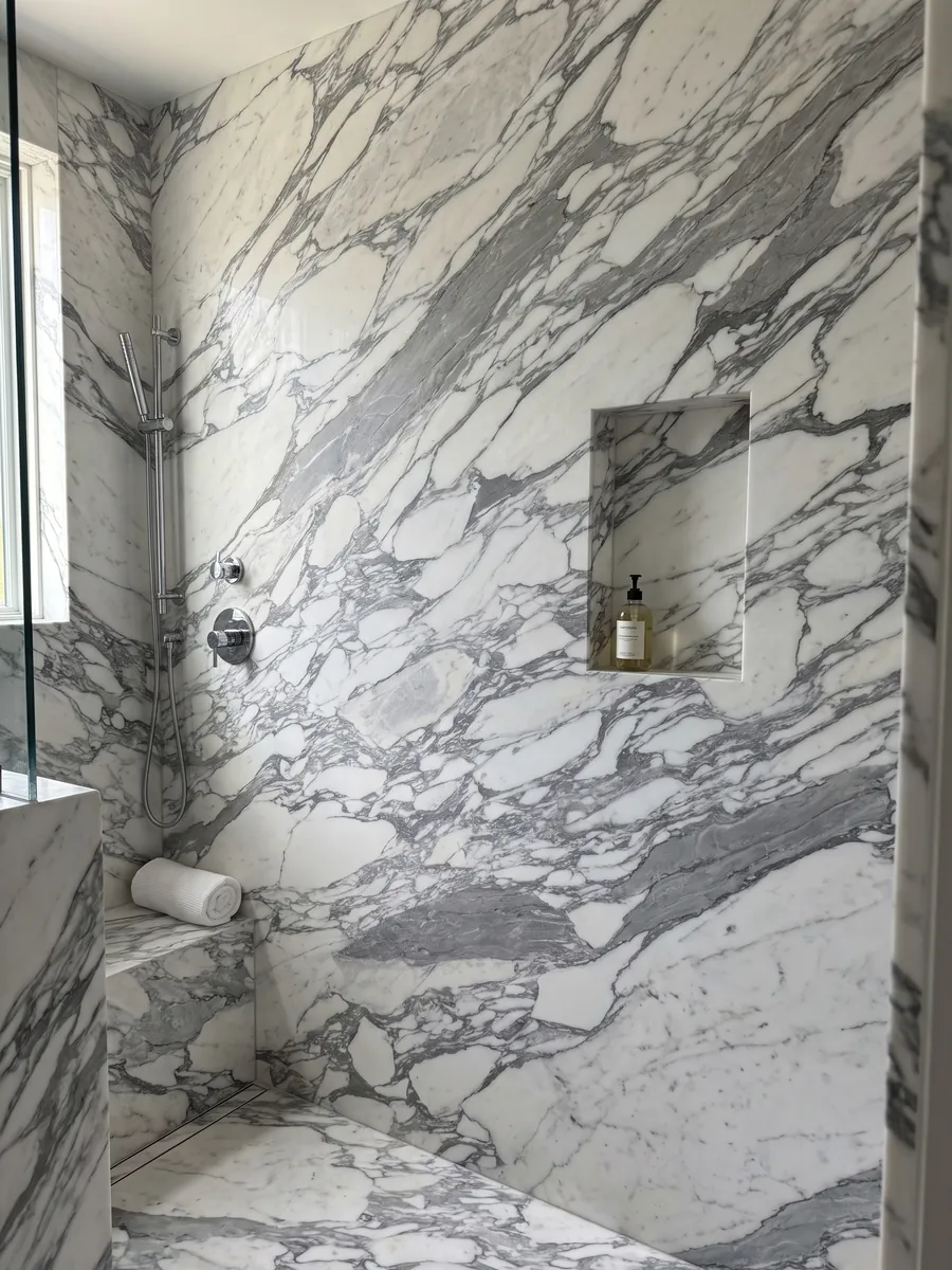Shower clad in whole slabs of white Arabesque marble with a recessed niche: elongated white fragments wrapped in thick gray veins running diagonally