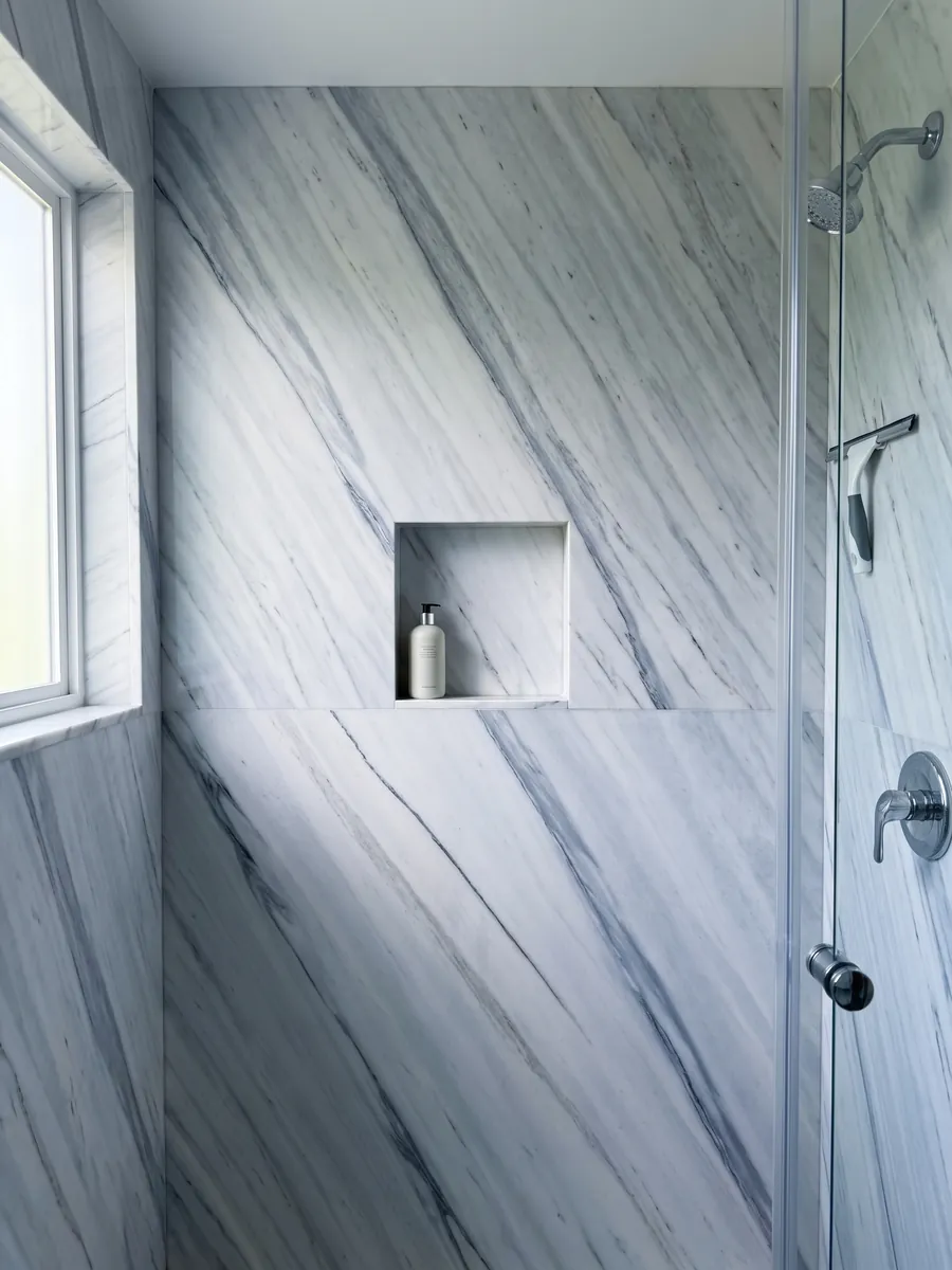 Shower clad in full slabs of white Bianco Covelano marble with a recessed niche: straight blue-gray veins, diagonal and parallel, over white