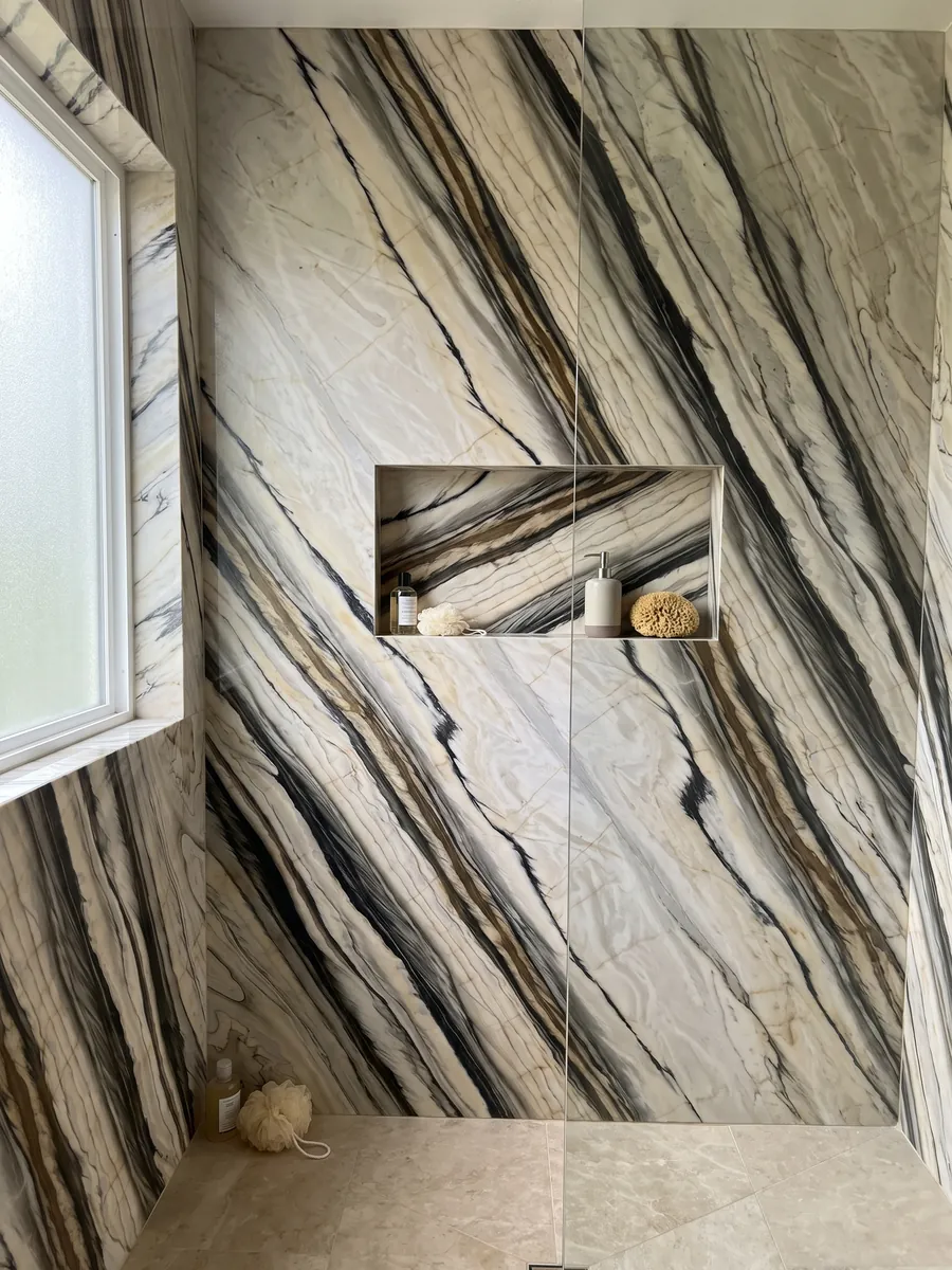 Shower clad in whole white Calacatta Oyster marble slabs with a recessed niche: wide black and brown bands running parallel in a diagonal over cream