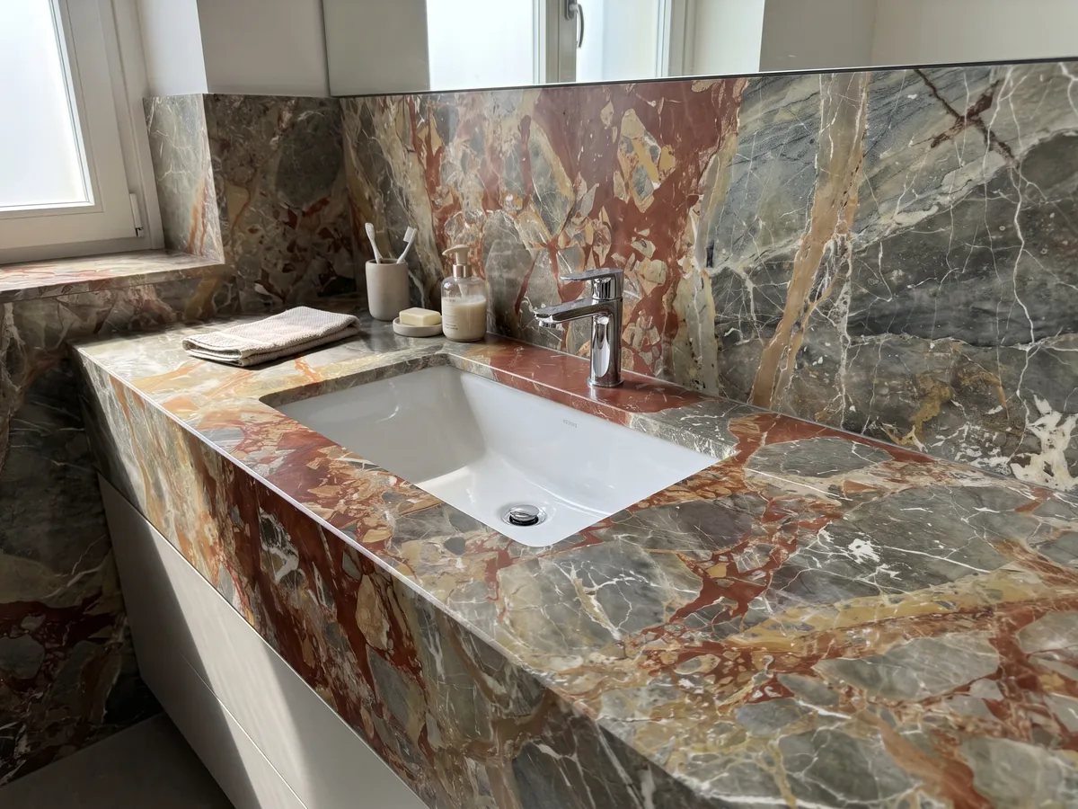 Bathroom vanity top and wall in gray Macchia Vecchia marble with a white sink and chrome faucet: greenish gray fragments with large rust-red patches and white veins