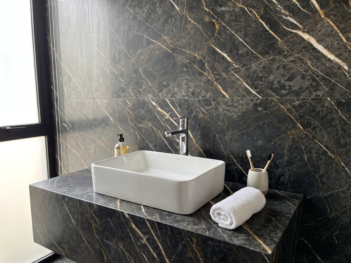 Bathroom vanity top and wall in black Port Laurent marble with a white sink and chrome fixtures: charcoal gray with gold and white veins running parallel in a diagonal