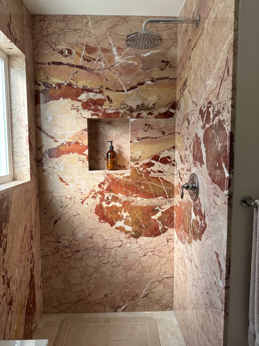 Shower clad in whole slabs of red Arlecchino marble with a recessed niche: large rust-red patches on pinkish beige, crossed by fine white veins