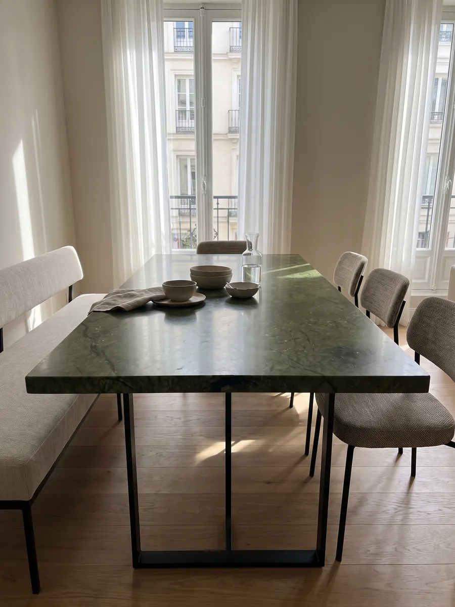 Dining table with a Verde Giada marble top on a steel base: intense olive green with tonal clouds and fine dark cracks; a dark blotch at the lower left