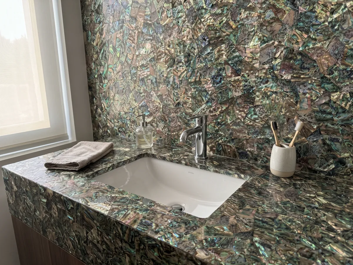 Vanity top and bathroom wall in abalone mother of pearl with a white sink and chrome faucets: a tight mosaic of iridescent shell in greens, blues and browns